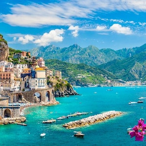 May include: Scenic coastal view of a town with colorful buildings, arched bridges, and a rocky cliffside. Turquoise water with boats and a mountain range under a blue sky with clouds. Pink flowers in the foreground.