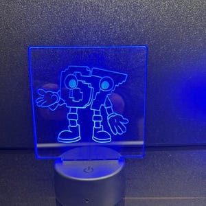 May include: A blue LED night light featuring a cartoon robot design. The robot has a square head with a plus sign, arms, and legs. The light sits on a black base with a power button.