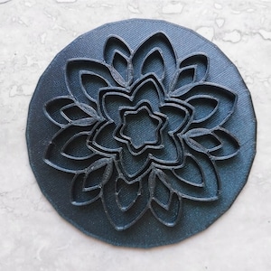 May include: A black circular cookie cutter featuring an intricate mandala design. The cutter has multiple layers of detailed patterns, creating a complex and symmetrical design.