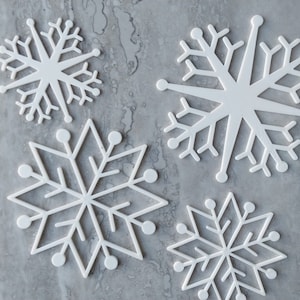 May include: Four white paper snowflakes of different sizes and designs on a grey textured background.