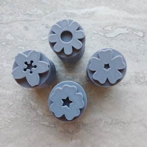 May include: Four gray plastic flower-shaped cookie cutters with different designs. The cutters are on a light gray marble surface.