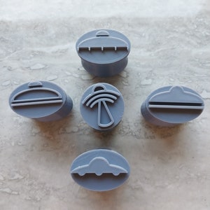 May include: Five grey plastic cookie cutters shaped like UFOs. The cutters have different designs, including a classic UFO shape, a UFO with a beam of light, and a UFO with a dome.