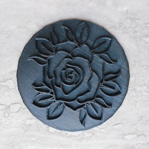 May include: A dark gray circular stamp with a raised design of a single rose with leaves.