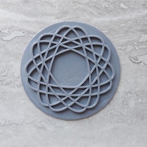 May include: A gray 3D printed coaster with a circular design featuring a geometric pattern of overlapping circles and lines.