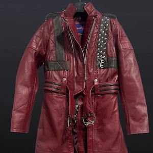 May include: A long, burgundy leather coat with black accents on the shoulders and upper chest. The coat features a zipper closure, studded details, and a belt with metal hardware. The coat is hanging on a black hanger.