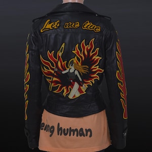 Op de afbeelding: Zwarte leren jas met geborduurd vlammen- en krijgerontwerp. De achterkant heeft de tekst "Let me live" boven een krijgersfiguur met een speer, omringd door vlammen. Vlammen sieren ook de mouwen.