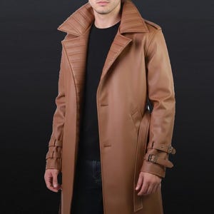 Pode incluir: Um trench coat de couro castanho claro com gola larga e fecho de botões. O casaco apresenta uma cintura com cinto e detalhes de fivelas nas mangas. O casaco é comprido até ao joelho e tem um design clássico.