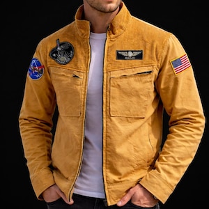 May include: Mustard yellow jacket with a corduroy collar and various patches. It has a zip closure, two chest pockets, and an American flag patch. The jacket is worn over a white t-shirt and paired with black jeans.