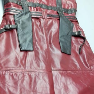 May include: A burgundy leather coat with black accents. The coat features a belted waist, shoulder straps, and decorative stitching. The leather has a glossy finish, and the coat appears to be new.