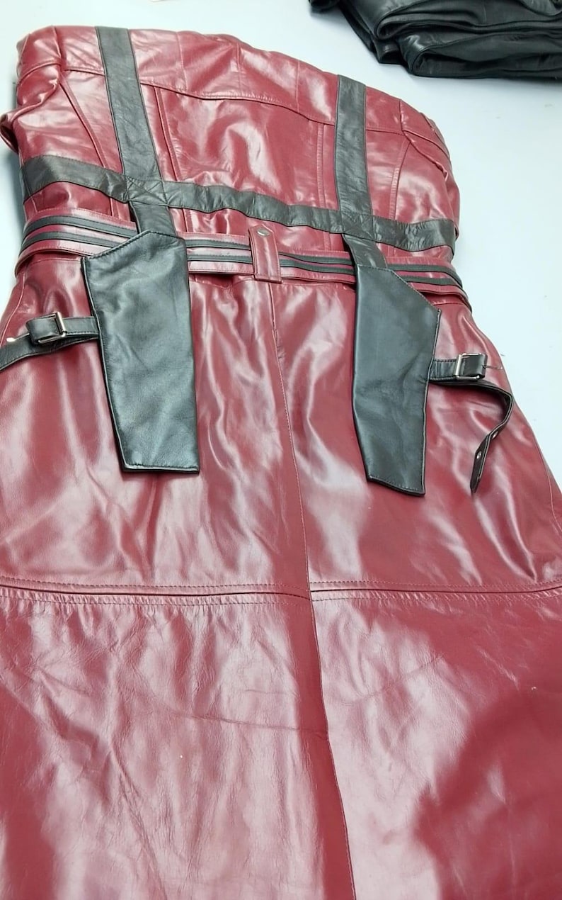 May include: A burgundy leather coat with black accents. The coat features a belted waist, shoulder straps, and decorative stitching. The leather has a glossy finish, and the coat appears to be new.