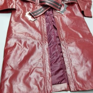 May include: A burgundy leather coat with a belted waist and black accents. The coat features a zipper closure, studded details, and a burgundy lining. The coat is a long length and has a classic, edgy style.