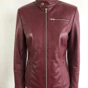 May include: A burgundy leather jacket with a stand-up collar and a silver zipper closure. The jacket has two zippered pockets on the front and a small pocket on the chest. The jacket is displayed on a mannequin.