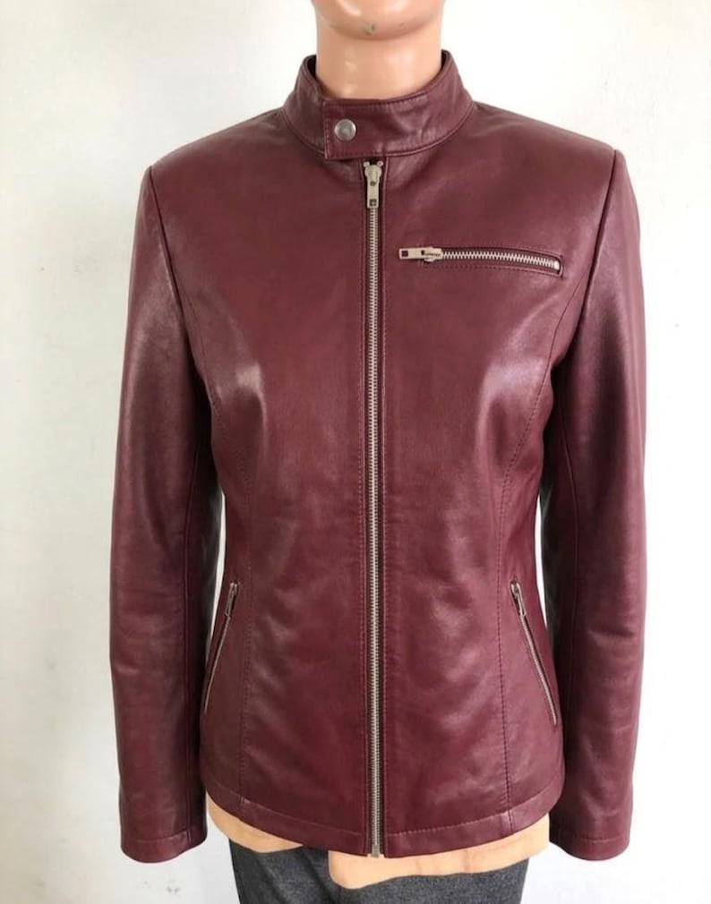 May include: A burgundy leather jacket with a stand-up collar and a silver zipper closure. The jacket has two zippered pockets on the front and a small pocket on the chest. The jacket is displayed on a mannequin.