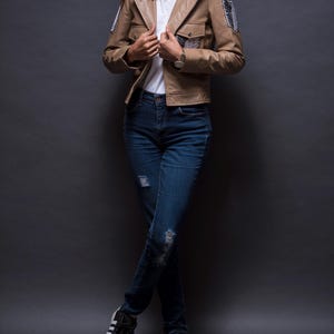 Może przedstawiać: Jasnobrązowa sk&oacute;rzana kurtka z dwiema przednimi kieszeniami i naszywkami na rękawach. Kurtka jest noszona na białym t-shircie i połączona z niebieskimi dżinsami oraz czarno-białymi trampkami.