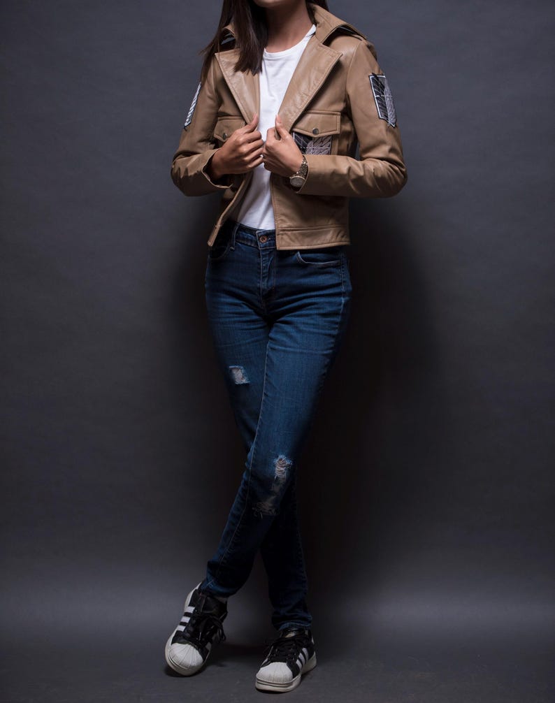 Może przedstawiać: Jasnobrązowa sk&oacute;rzana kurtka z dwiema przednimi kieszeniami i naszywkami na rękawach. Kurtka jest noszona na białym t-shircie i połączona z niebieskimi dżinsami oraz czarno-białymi trampkami.