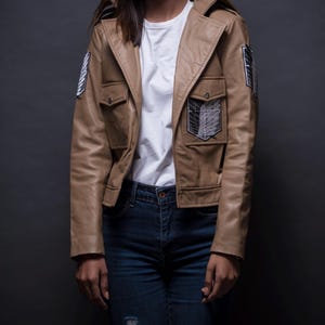 Może przedstawiać: Jasnobrązowa kurtka ze sztucznej sk&oacute;ry z dwiema przednimi kieszeniami i kołnierzem z klapami. Kurtka posiada haftowane naszywki na rękawach i piersi. Osoba ma na sobie biały t-shirt i niebieskie dżinsy.