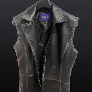 Könnte beinhalten: Eine schwarze Lederweste mit Reverskragen und geflochtenem Besatz an den Kanten. Die Weste hat eine taillierte Passform und eine Schnürung vorne. Das Etikett "Iman Zah" ist innen sichtbar.