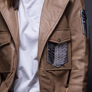 Może przedstawiać: Jasnobrązowa sk&oacute;rzana kurtka z dwiema przednimi kieszeniami i czarno-szarą naszywką z emblematem na piersi. Kurtka ma kołnierz i jest noszona na białym t-shircie i niebieskich dżinsach.