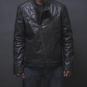 Pode incluir: Casaco de couro preto com zíper assimétrico, detalhes acolchoados nos ombros e um pequeno emblema no peito esquerdo. O casaco tem gola alta e corte ajustado. O modelo usa jeans azuis.