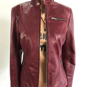 May include: A burgundy leather jacket with a stand-up collar and a silver zipper closure. The jacket has two zippered pockets and a tailored fit. The jacket is shown on a mannequin.