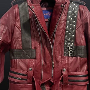 May include: A burgundy leather jacket with black accents and silver stud details. The jacket features a zipper closure, a collar, and a belt. The shoulder straps are black with silver studs. The jacket has the brand name "Iman" on the label.