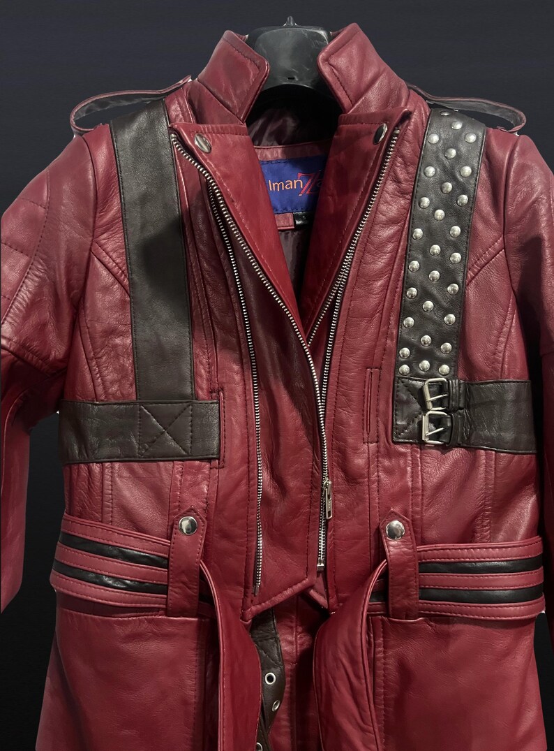 May include: A burgundy leather jacket with black accents and silver stud details. The jacket features a zipper closure, a collar, and a belt. The shoulder straps are black with silver studs. The jacket has the brand name "Iman" on the label.