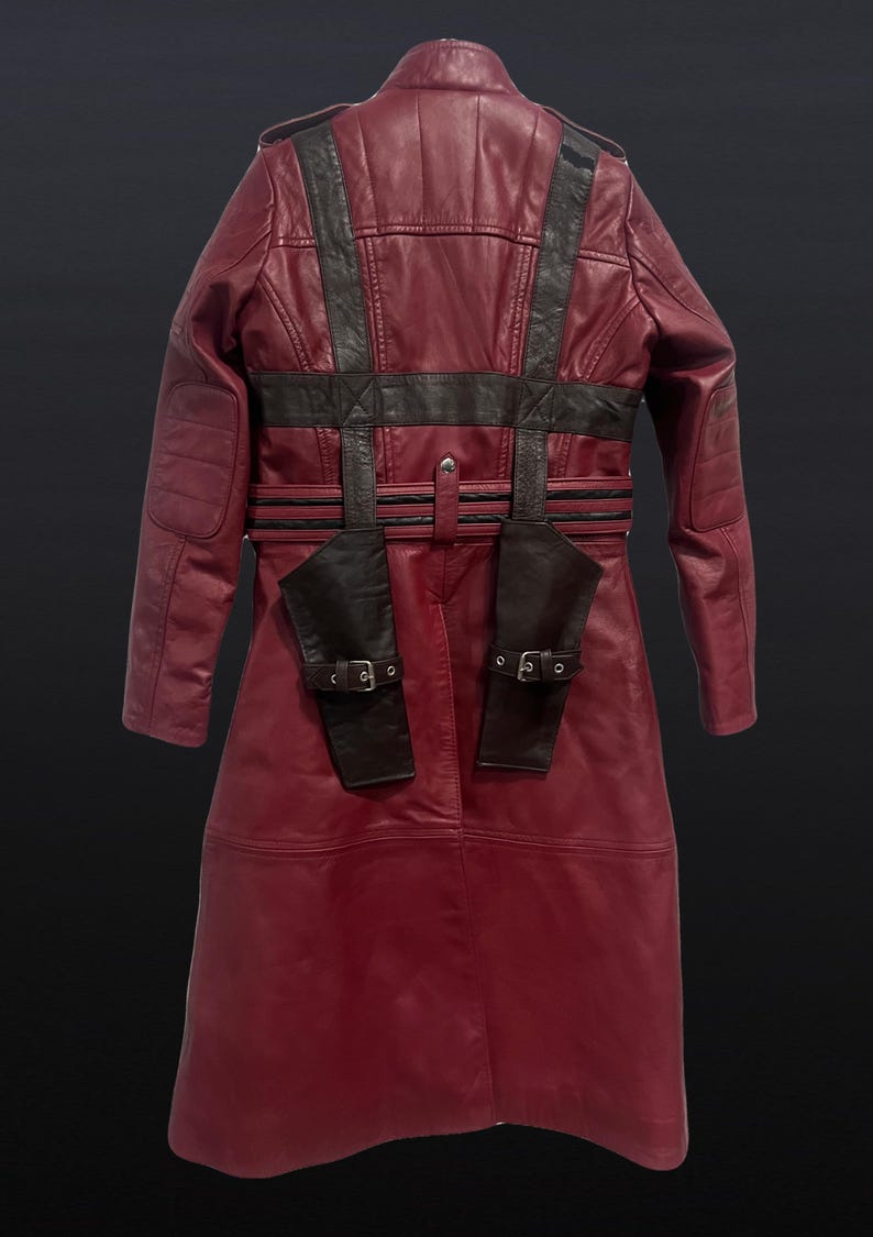 May include: A burgundy leather coat with black accents. The coat features a long design with a belted waist, shoulder straps, and two small pockets with buckles. The back of the coat is visible, showcasing the detailed stitching and design.