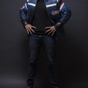Puede incluir: Chaqueta de cuero azul acolchada con rayas blancas y rojas en las mangas y hombros. La chaqueta tiene cuello alto y un parche en el pecho. La persona lleva una camiseta negra, vaqueros azul oscuro y zapatillas rojas.