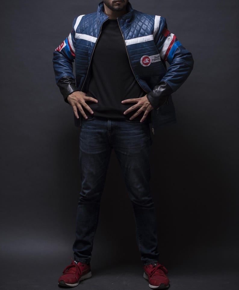 Puede incluir: Chaqueta de cuero azul acolchada con rayas blancas y rojas en las mangas y hombros. La chaqueta tiene cuello alto y un parche en el pecho. La persona lleva una camiseta negra, vaqueros azul oscuro y zapatillas rojas.