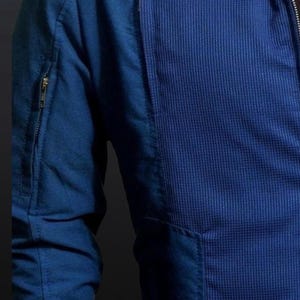 May include: Close-up of a blue jacket with a textured waffle-weave panel on the front. The jacket features a zippered sleeve pocket and a front pocket. The fabric appears to be a medium-weight material, suitable for cooler weather.