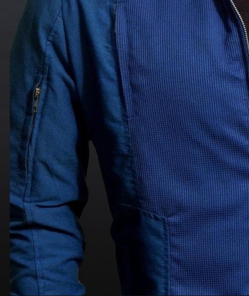 May include: Close-up of a blue jacket with a textured waffle-weave panel on the front. The jacket features a zippered sleeve pocket and a front pocket. The fabric appears to be a medium-weight material, suitable for cooler weather.