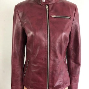 May include: A burgundy leather jacket with a stand-up collar and a silver zipper closure. The jacket has two zippered pockets on the front and a small zippered pocket on the chest. The sleeves are long and fitted.
