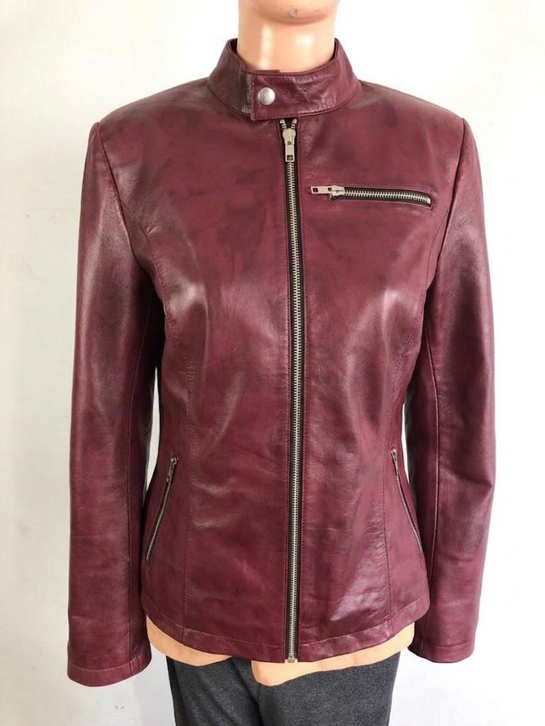 May include: A burgundy leather jacket with a stand-up collar and a silver zipper closure. The jacket has two zippered pockets on the front and a small zippered pocket on the chest. The sleeves are long and fitted.