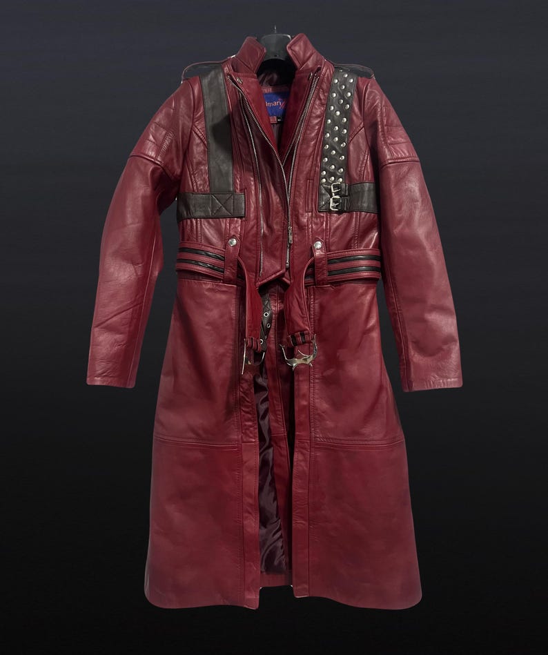 May include: A long, burgundy leather coat with black accents on the shoulders and upper chest. The coat features a zipper closure, studded details, and a belt with metal hardware. The coat is hanging on a black hanger.