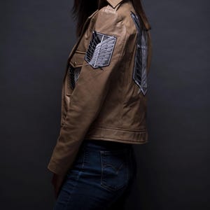 Może przedstawiać: Jasnobrązowa sk&oacute;rzana kurtka z czarno-białymi haftowanymi naszywkami na ramieniu i plecach. Kurtka ma kołnierz, zapinane na guziki kieszenie i dopasowany kr&oacute;j. Osoba ma na sobie niebieskie dżinsy.