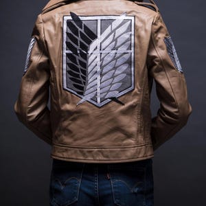 Może przedstawiać: Brązowa sk&oacute;rzana kurtka z dużym emblematem na plecach, przedstawiającym czarne, szare i białe skrzydła w prostokątnej ramie. Kurtka ma klasyczny kołnierz i mniejsze emblematy na rękawach. Osoba ma na sobie niebieskie dżinsy.