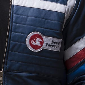 Puede incluir: Chaqueta de cuero azul marino con detalles en blanco y rojo. La chaqueta presenta un dise&ntilde;o acolchado y un parche con la inscripci&oacute;n "Dead Pegasus" y un logotipo rojo y blanco. Una cremallera recorre la parte delantera.