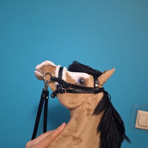 Op de afbeelding: Een pluche paardenhoofd in tinten bruin en crème, met een zwarte manen en een zwart halster. Het paard heeft grote ogen en wordt door een hand tegen een blauwe achtergrond gehouden.