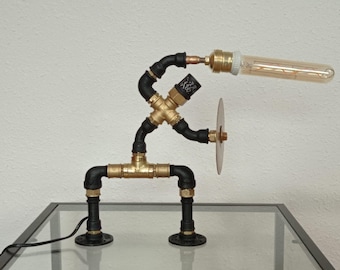 May include: A unique industrial-style desk lamp crafted from black and brass pipes, resembling a stick figure. The lamp features an Edison-style bulb and a small, circular metal accent. It sits on a glass surface.