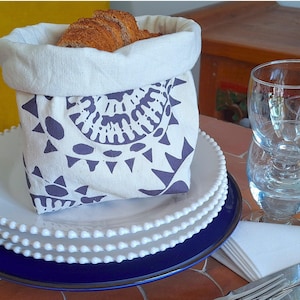Peut inclure: Un sac à pain blanc et violet rempli de pain tranché, posé sur une pile d'assiettes blanches à bord bleu. Des verres transparents et des couverts sont également visibles, suggérant un cadre de restauration.