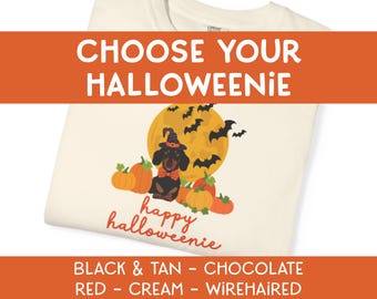 May include: A cream-colored t-shirt featuring a graphic of a dachshund wearing a witch hat and orange bowtie, surrounded by pumpkins and bats against a yellow moon. The text reads CHOOSE YOUR HALLOWEENIE, happy halloweenie, and BLACK & TAN - CHOCOLATE RED - CREAM - WIREHAIRED.