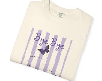 May include: A cream-colored t-shirt featuring a graphic design with vertical light purple stripes. The text "Bye Bye" is printed in a purple script font above a purple butterfly illustration with a dashed black line trailing behind it.