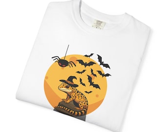 May include: A white t-shirt featuring a graphic of a spotted gecko wearing a black witch hat, perched on a stone. The background shows a large yellow moon, a black spider hanging from a thread, and a flock of black bats flying across the moon.