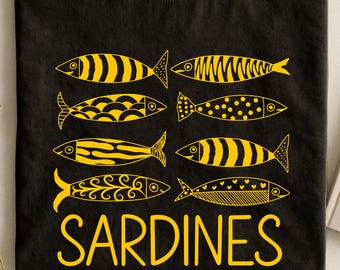 May include: A black t-shirt featuring a graphic print of eight stylized fish arranged in two columns. Each fish is colored in bright yellow and displays a unique pattern, including stripes, dots, and swirls. The word SARDINES is printed in yellow.