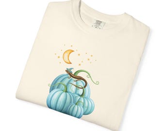 May include: A cream-colored t-shirt featuring a graphic of a brown lizard wearing a blue wizard hat perched on a cluster of light blue pumpkins. Above the pumpkins, a yellow crescent moon and several small yellow stars are illustrated.