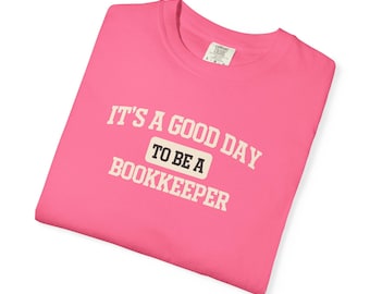May include: A bright pink t-shirt with the text "IT'S A GOOD DAY TO BE A BOOKKEEPER" in white block letters. The text is centered on the front of the shirt. The shirt is folded and appears to be made of a soft material.