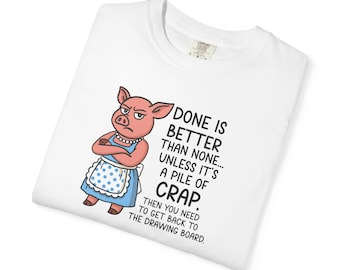 May include: A white t-shirt featuring a cartoon pig wearing a blue apron with white polka dots and a pearl necklace. The pig has crossed arms and a stern expression. The text reads: DONE IS BETTER THAN NONE... UNLESS IT'S A PILE OF CRAP. THEN YOU NEED TO GET BACK TO THE DRAWING BOARD.