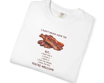 May include: A white t-shirt featuring a graphic of two strips of cooked bacon in the center. Above the bacon is the text I DON'T BRING HOME THE and below it is the text BUT... I SHOP FOR IT. I COOK IT. I SERVE IT. I SAVE THE GREASE. I WASH THE PAN. YOU'RE WELCOME.