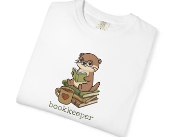 May include: A white t-shirt featuring a graphic of a brown otter wearing glasses, sitting on a stack of books while reading a green book. Next to the books is a brown mug with a heart design. The text "bookkeeper" is printed in green below.