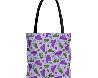 May include: A tote bag with a light purple background featuring a repeating pattern of green and purple frogs, small mushrooms in shades of blue and green, and tiny dark blue star-like shapes. The bag has two black fabric shoulder straps.
